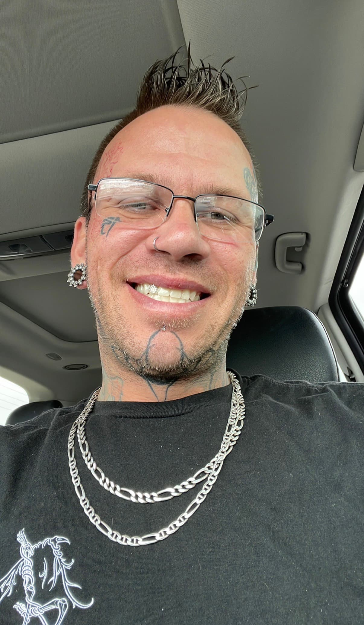 Smiling man with spiked hair, facial tattoos, glasses, and silver chains sitting in a car.