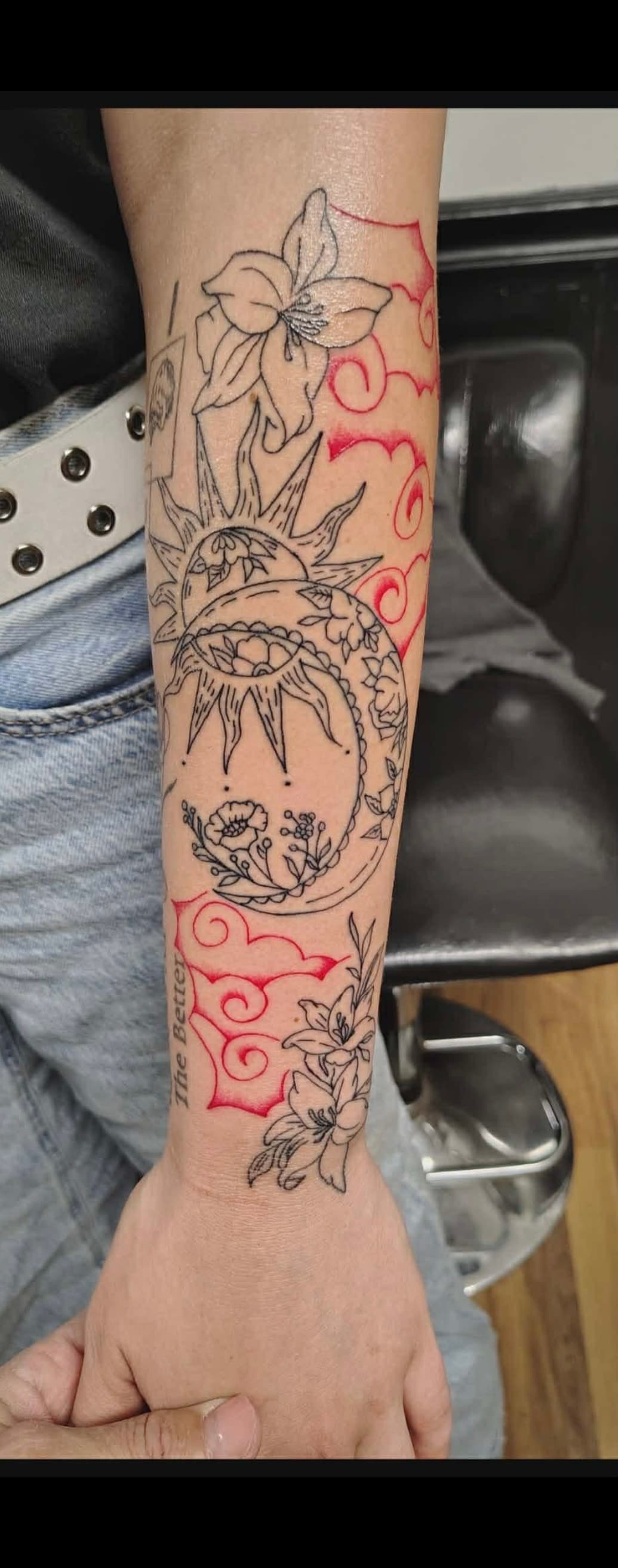 Forearm tattoo featuring black ink sun, moon, and lilies with red stylized cloud accents.