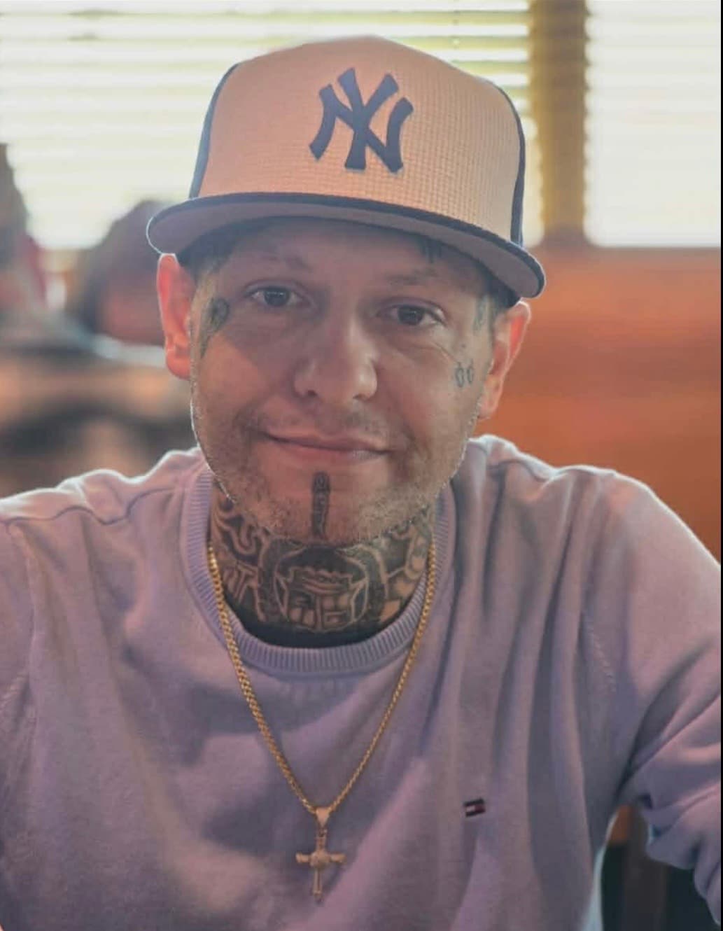 Smiling man with face and neck tattoos wearing a Yankees cap and gold cross necklace.