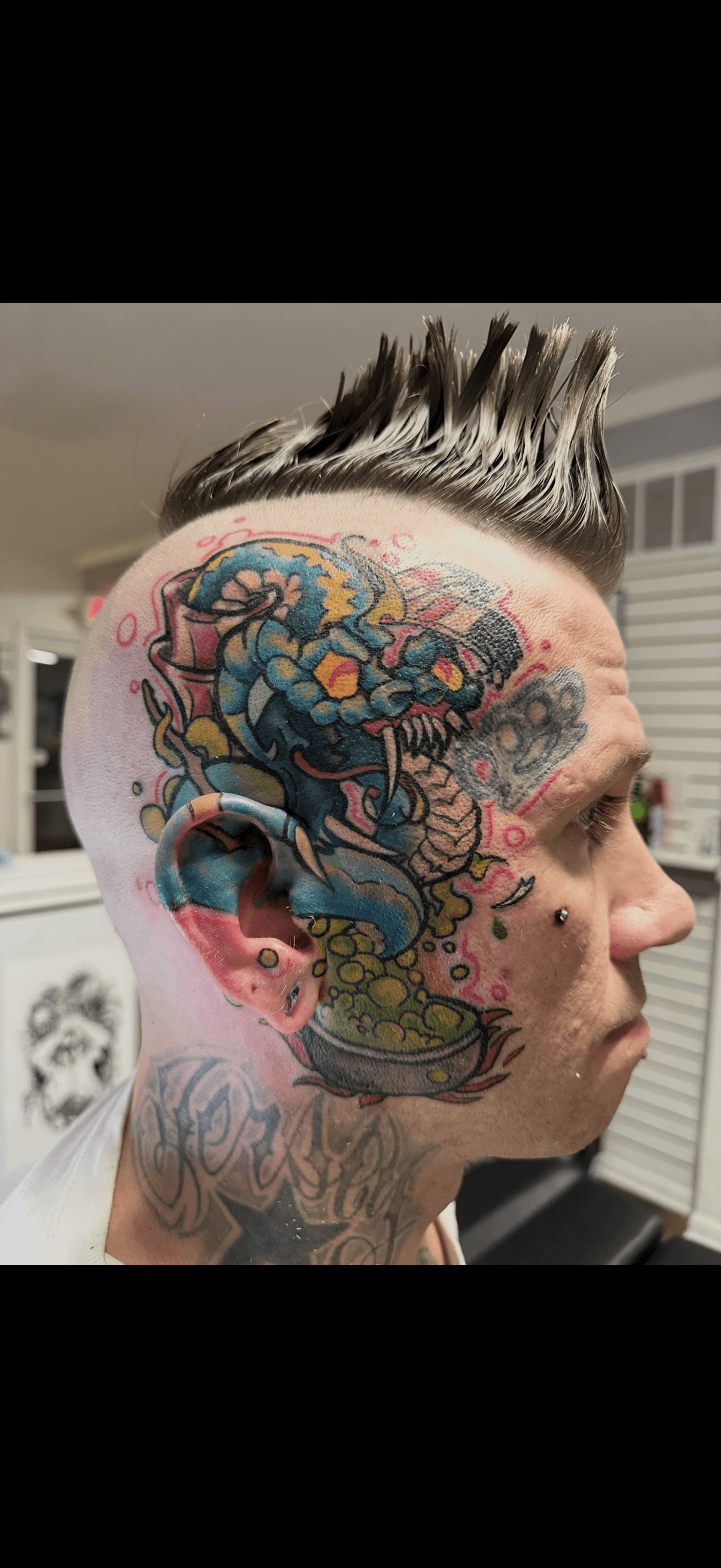 Man with a mohawk and a vibrant monster tattoo covering his head and ear.