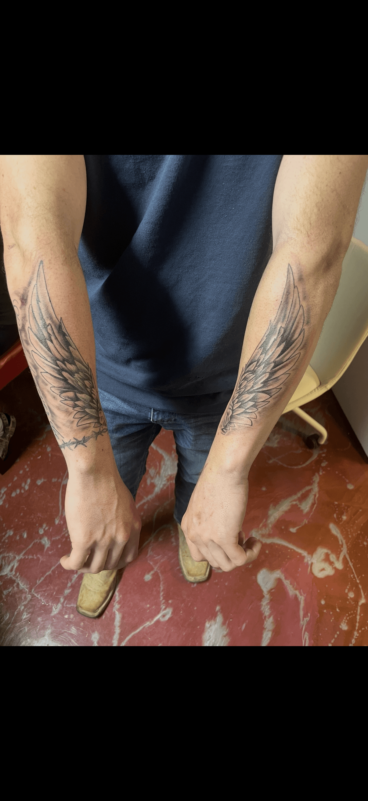 Detailed black and grey feathered wing tattoos on both forearms, with a barbed wire accent.