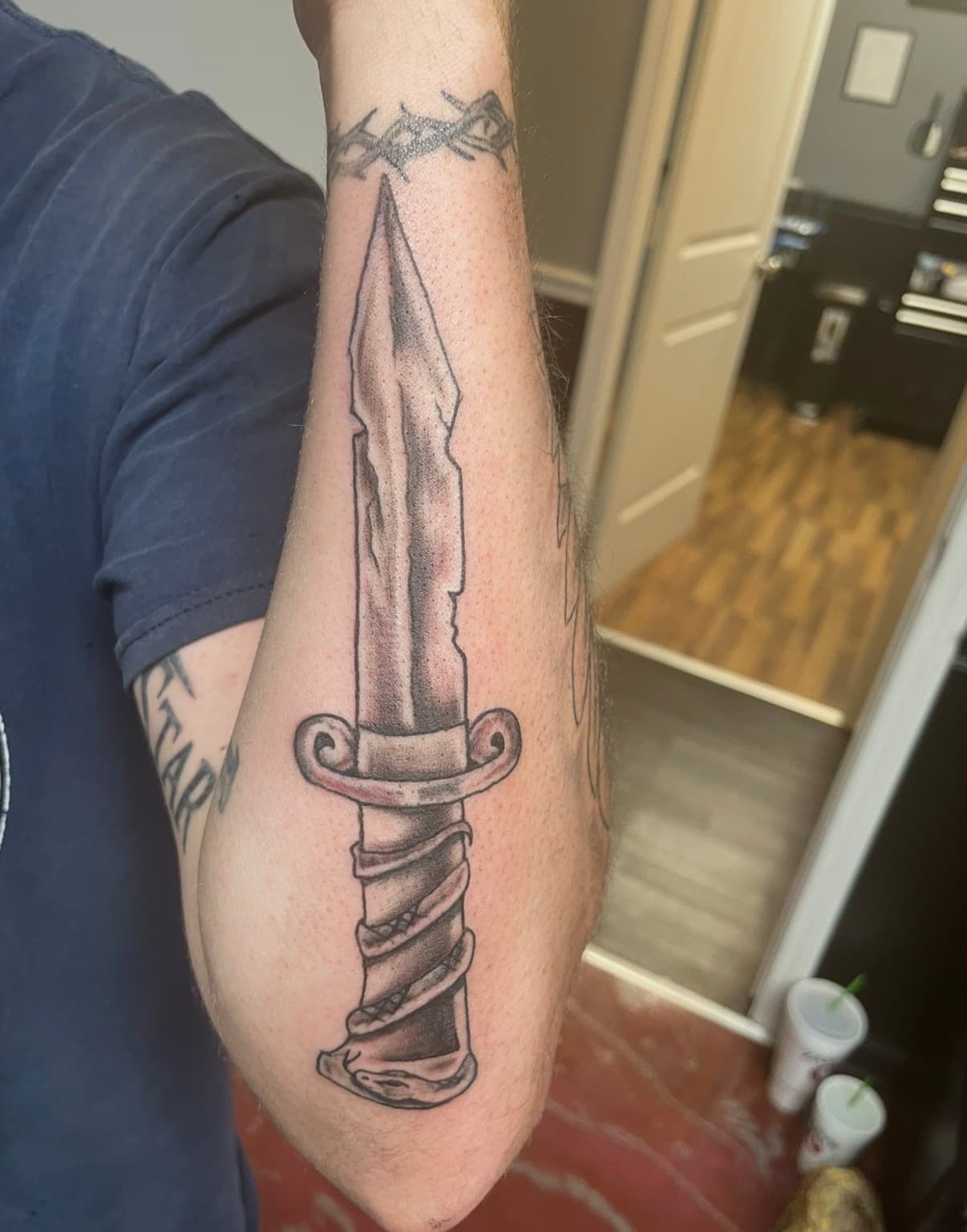Black and grey tattoo of a chipped dagger with an ornate hilt on a forearm.