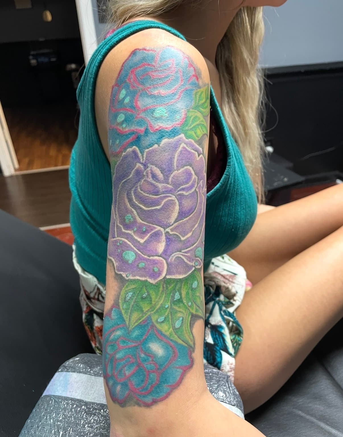 Colorful tattoo of purple and blue roses with green leaves and water droplets on an arm.