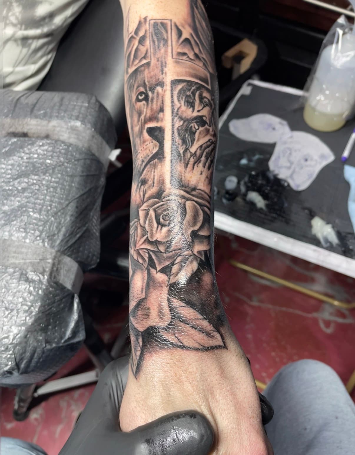 Detailed black and grey forearm tattoo featuring a cross, lion, praying figure, and a rose.