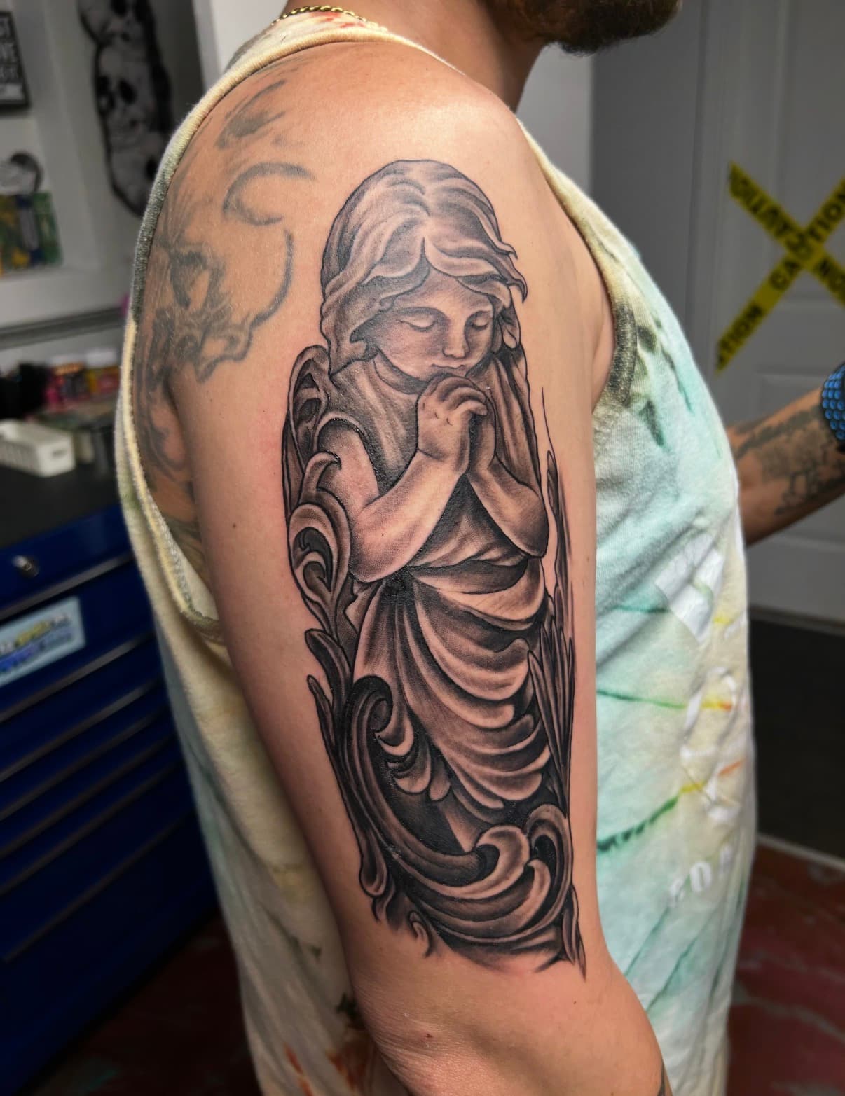 Detailed black and grey tattoo of a praying angel with folded hands on an upper arm.